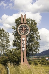 A tall wooden sculpture set in a rural landscape features a circular emblem with intricate geometric patterns at the top. Below the emblem, a vertical structure displays stylized text. The sculpture is surrounded by tall grasses and trees, with a backdrop of rolling hills and scattered houses under a partly cloudy sky.
