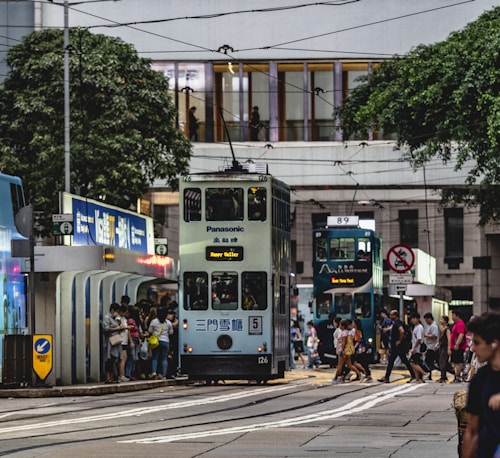 A busy urban scene featuring two double-decker trams on parallel tracks in a bustling city environment. People are waiting at a tram stop and crossing the street, surrounded by buildings and lush green trees.