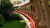 A red pipeline runs alongside a series of evenly spaced pillars, bordered by green grass and lush vegetation. The scene includes a walkway that follows the curvature of the pipeline. Buildings and foliage are visible in the background.