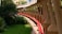 A red pipeline runs alongside a series of evenly spaced pillars, bordered by green grass and lush vegetation. The scene includes a walkway that follows the curvature of the pipeline. Buildings and foliage are visible in the background.