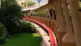 A red pipeline runs alongside a series of evenly spaced pillars, bordered by green grass and lush vegetation. The scene includes a walkway that follows the curvature of the pipeline. Buildings and foliage are visible in the background.
