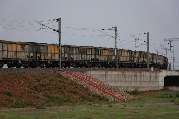 A long freight train is seen running on a track elevated over a concrete structure. The carriages of the train are cargo containers with visible signs of wear such as rust and dirt. The train is supported by electrical poles and wires running parallel to it. The surrounding area features a mix of grass, vegetation, and exposed soil. There is a small, stepped embankment leading up to the track, indicated by a red and white stripe.
