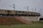 A long freight train is seen running on a track elevated over a concrete structure. The carriages of the train are cargo containers with visible signs of wear such as rust and dirt. The train is supported by electrical poles and wires running parallel to it. The surrounding area features a mix of grass, vegetation, and exposed soil. There is a small, stepped embankment leading up to the track, indicated by a red and white stripe.