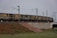 A long freight train is seen running on a track elevated over a concrete structure. The carriages of the train are cargo containers with visible signs of wear such as rust and dirt. The train is supported by electrical poles and wires running parallel to it. The surrounding area features a mix of grass, vegetation, and exposed soil. There is a small, stepped embankment leading up to the track, indicated by a red and white stripe.