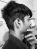 A black and white profile view of a person with thick, styled hair. The person is touching their chin thoughtfully, conveying a pensive mood. The background is a simple indoor setting with visible walls and some indistinct items.