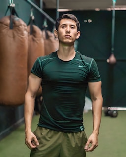 A person is standing confidently in a gym-like setting, wearing a dark green athletic shirt and light brown shorts. Several punching bags are visible hanging in the background, suggesting a boxing or fitness environment.