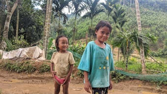 Two young children are standing outside on a dirt path. They are surrounded by lush green vegetation and tall trees. The child on the left is wearing a beige shirt and pink shorts, while the child on the right is in a blue shirt with a cactus design. Both children have happy expressions and appear to be enjoying their natural surroundings.