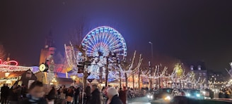 A large, brightly lit Ferris wheel dominates the center, surrounded by festive Christmas market stalls with decorative lights. People walk along the street, creating a lively atmosphere. The background includes illuminated trees and a tall building with a clock.