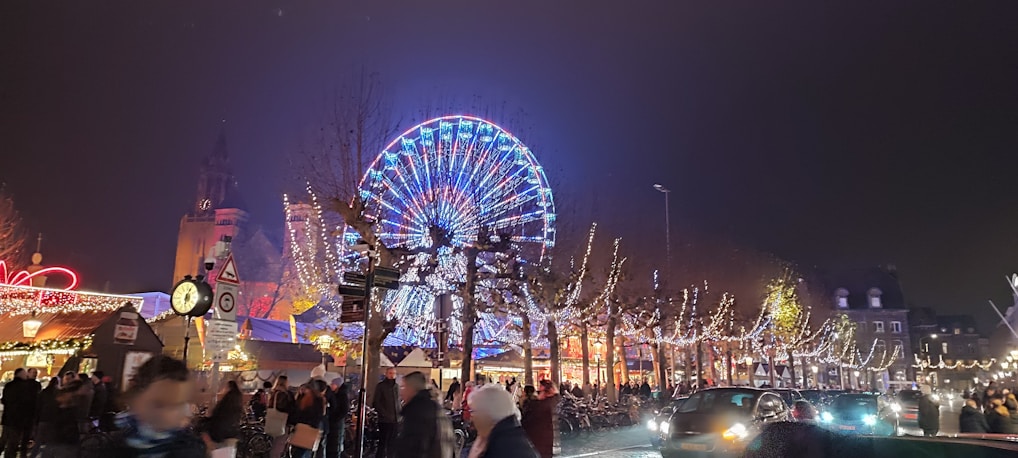 A large, brightly lit Ferris wheel dominates the center, surrounded by festive Christmas market stalls with decorative lights. People walk along the street, creating a lively atmosphere. The background includes illuminated trees and a tall building with a clock.