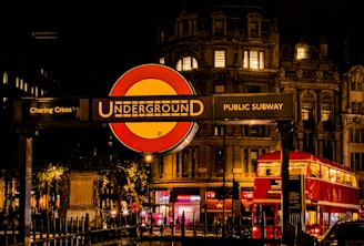 A nighttime scene featuring a prominent London Underground sign at Charing Cross. The background includes an illuminated historic building and a classic red double-decker bus driving by. Ambient city lights and dark sky set a lively yet serene urban atmosphere.