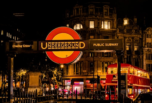A nighttime scene featuring a prominent London Underground sign at Charing Cross. The background includes an illuminated historic building and a classic red double-decker bus driving by. Ambient city lights and dark sky set a lively yet serene urban atmosphere.