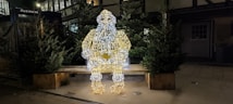A large light sculpture of Santa Claus made from wire and illuminated with white and yellow lights is placed on a wooden bench. The setting is outdoors, surrounded by several green pine trees in wooden planters, suggesting a festive or holiday theme. The background includes a partially visible building with windows.