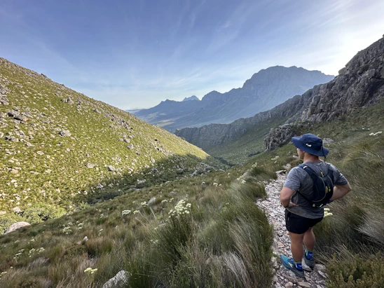 A smiling traveler standing on a scenic mountain trail in Georgia, surrounded by lush greenery and blue skies.