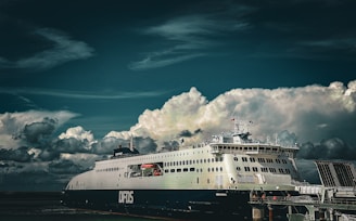 A large ferry is docked at a port under a dramatic sky filled with thick, white clouds. The ferry is prominently labeled DFDS, and appears well-maintained, suggesting it might be used for passenger or vehicle transportation.