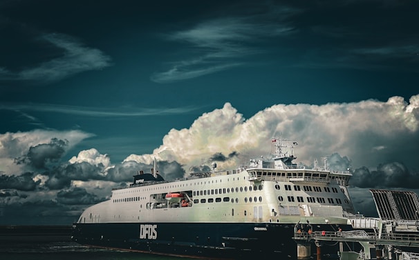 A large ferry is docked at a port under a dramatic sky filled with thick, white clouds. The ferry is prominently labeled DFDS, and appears well-maintained, suggesting it might be used for passenger or vehicle transportation.