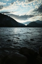 A tranquil lakeside scene during sunset or sunrise with gentle ripples on the water's surface. Dark silhouettes of mountains frame the horizon, where the sun casts a glow through scattered clouds.