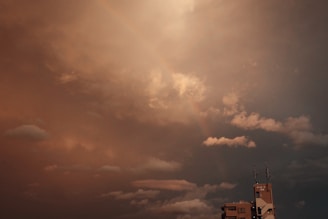 A dramatic sky with dense, dark clouds and a faint, arching rainbow. At the bottom right corner, a building with antennas peeks into the frame, adding an urban element to the foreboding weather scene.