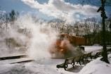 An orange truck is emitting steam or smoke in a snowy park-like setting. The area is surrounded by barren trees and benches are visible on either side. The sky is partly cloudy, casting a soft winter light over the scene.
