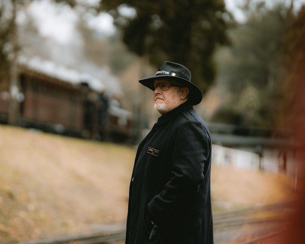 Un hombre con un abrigo negro y sombrero parado en una vía de tren
