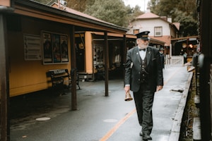 A railway station setting featuring a man in a station master's uniform walking down the wet platform. He carries a bell in his hand. The platform is lined with wooden buildings and benches with 'Belgrave' signage. Posters are displayed on the walls, and the background shows trees and a red-roofed building.