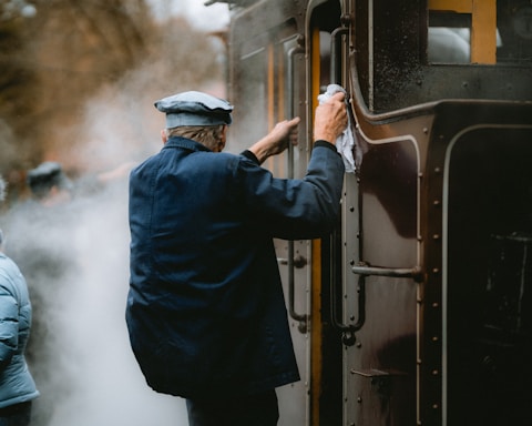 An older person in a blue uniform and cap is cleaning the exterior of a vintage train, using a cloth. The scene is enveloped in steam, suggesting the train is in operation. Another person in a blue jacket is visible, partially obscured by the steam.