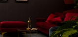 A sophisticated, dimly lit interior with a luxurious red velvet sofa and matching ottoman set against a dark wall. A small, round coffee table stands in front with a golden bird-shaped figurine and a few papers. Lush green plants add a touch of nature on the right side of the image.