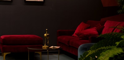 A sophisticated, dimly lit interior with a luxurious red velvet sofa and matching ottoman set against a dark wall. A small, round coffee table stands in front with a golden bird-shaped figurine and a few papers. Lush green plants add a touch of nature on the right side of the image.
