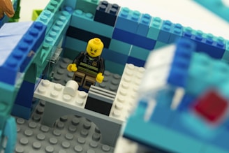 A LEGO figure with a yellow face and an intense expression stands inside a structure built from blue and white LEGO bricks. The figure is placed in a room with walls made from alternating shades of blue tiles. A small white table with a few blocks on it is in front of the figure.