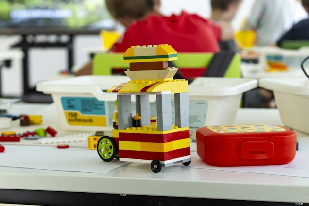 A small LEGO model resembling a food cart is placed on a table alongside scattered LEGOs and containers. In the background, several people are seated, partially out of focus. The cart features bright colors including yellow, red, and green, with a distinct umbrella-like top.