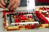 Two hands are assembling a structure using various colored LEGO bricks. The structure appears to be partially built on a grey baseplate, with colors including red, black, white, brown, and yellow. Various brick shapes and sizes are scattered around the area.