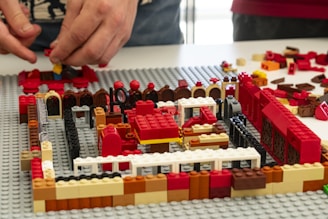 Two hands are assembling a structure using various colored LEGO bricks. The structure appears to be partially built on a grey baseplate, with colors including red, black, white, brown, and yellow. Various brick shapes and sizes are scattered around the area.