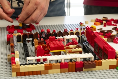 Two hands are assembling a structure using various colored LEGO bricks. The structure appears to be partially built on a grey baseplate, with colors including red, black, white, brown, and yellow. Various brick shapes and sizes are scattered around the area.