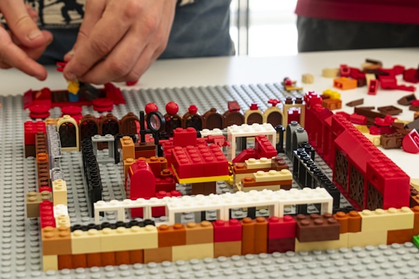 Two hands are assembling a structure using various colored LEGO bricks. The structure appears to be partially built on a grey baseplate, with colors including red, black, white, brown, and yellow. Various brick shapes and sizes are scattered around the area.