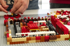 Two hands are assembling a structure using various colored LEGO bricks. The structure appears to be partially built on a grey baseplate, with colors including red, black, white, brown, and yellow. Various brick shapes and sizes are scattered around the area.