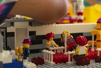 A close-up of a child's arm hovering over a colorful LEGO scene depicting several minifigures interacting in a playful setting. The scene includes small fences, a red floor, and various LEGO bricks forming structures around the figures.