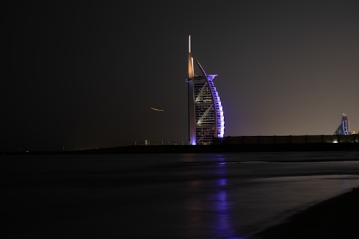 A prominent, sail-shaped skyscraper illuminated at night against a dark sky, with a smooth reflection on the nearby water surface. The building's lights feature purples and whites, creating a striking visual contrast.