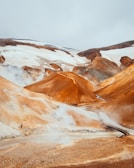 A dramatic landscape featuring geothermal steam vents and rolling hills with reddish-brown and white snow patches contrasting against the sky. A small wooden bridge crosses a narrow river.