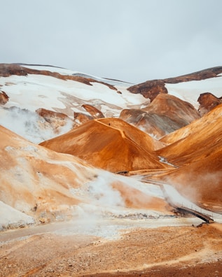 A dramatic landscape featuring geothermal steam vents and rolling hills with reddish-brown and white snow patches contrasting against the sky. A small wooden bridge crosses a narrow river.