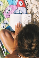 A person with braided hair sits on a towel with colorful cartoon faces, reading a book. The surrounding area is rocky, suggesting a beach setting. The person's nails are painted white and they are wearing a thin bracelet.