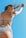 A person wearing a black and white striped bikini is captured from a low angle against a clear blue sky. The posture suggests movement, with arms raised and hair flowing slightly in the wind. Sunglasses add a stylish touch to the scene.