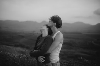A black and white photograph of two people in an intimate embrace against a blurred landscape backdrop. One person is resting their head back against the other's chest, eyes closed, while the other leans in with a serene expression. The scene conveys a sense of calm and connection.