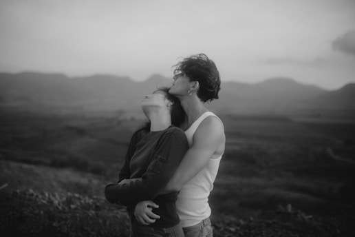 A black and white photograph of two people in an intimate embrace against a blurred landscape backdrop. One person is resting their head back against the other's chest, eyes closed, while the other leans in with a serene expression. The scene conveys a sense of calm and connection.