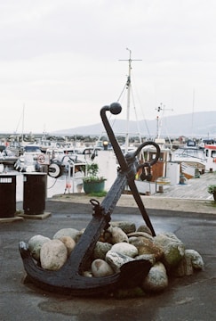 An anchor is placed on a pile of rocks near a harbor, surrounded by numerous boats and a clear sky in the background. The setting conveys a maritime atmosphere with various fishing or recreational vessels docked nearby.