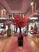A beautifully decorated interior features a striking centerpiece of a pink cherry blossom tree. The floor is adorned with soft pastel colors, and various decorative items such as plates and cloths are displayed on tables along the sides. Hanging from the ceiling are white floral garlands. Large windows allow natural light to fill the space, enhancing its inviting atmosphere.