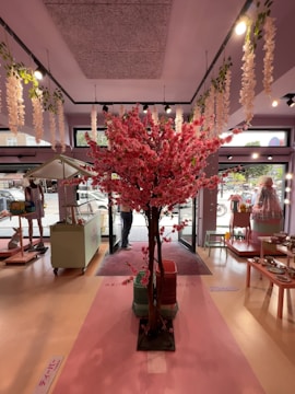A beautifully decorated interior features a striking centerpiece of a pink cherry blossom tree. The floor is adorned with soft pastel colors, and various decorative items such as plates and cloths are displayed on tables along the sides. Hanging from the ceiling are white floral garlands. Large windows allow natural light to fill the space, enhancing its inviting atmosphere.