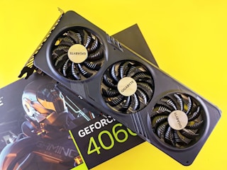 A graphics card with three fans prominently displaying the Gigabyte logo is positioned atop its packaging. The packaging, partly visible, features 'GeForce 4060' branding and a futuristic robotic design. The bright yellow background enhances the contrast of the black graphics card.