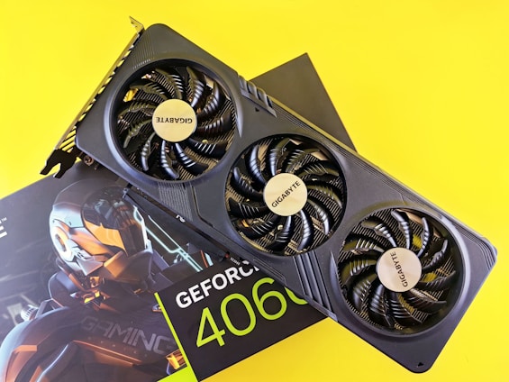 A graphics card with three fans prominently displaying the Gigabyte logo is positioned atop its packaging. The packaging, partly visible, features 'GeForce 4060' branding and a futuristic robotic design. The bright yellow background enhances the contrast of the black graphics card.