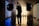 A silhouetted person stands in a photography studio, framed by two large studio lights on stands. The room is dimly lit with linear blue and yellow lights creating a contrasting background. Smoke or fog adds an atmospheric effect, with the floor reflecting the lights.