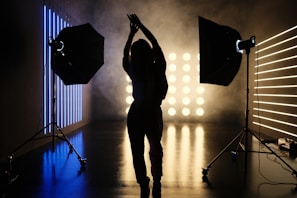 A silhouetted person stands in a photography studio, framed by two large studio lights on stands. The room is dimly lit with linear blue and yellow lights creating a contrasting background. Smoke or fog adds an atmospheric effect, with the floor reflecting the lights.