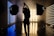 A silhouetted person stands in a photography studio, framed by two large studio lights on stands. The room is dimly lit with linear blue and yellow lights creating a contrasting background. Smoke or fog adds an atmospheric effect, with the floor reflecting the lights.